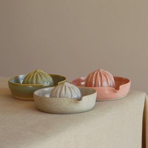 Puede incluir: Tres exprimidores de cítricos de cerámica en tonos verde, rosa y beige. Cada exprimidor tiene un escariador acanalado y un cuenco con boquilla. Los exprimidores se exhiben sobre un mantel de color neutro.