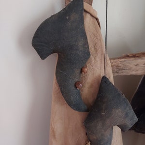 May include: Two dark gray felt elf shoes with bell accents hanging from black string. The shoes are a muted gray-green color and have a rustic, aged appearance. They are hanging in front of a tan fabric.