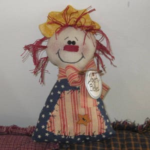 May include: A handmade rag doll with red hair, a yellow hat, and a red, white, and blue striped dress. The doll is holding a tag that says "Liberty Belle".