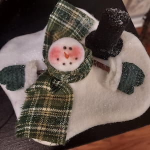 May include: A melting snowman with a green plaid scarf, black top hat, and black buttons for eyes. The snowman is on a white, glittery surface.
