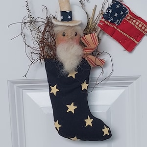 May include: A blue stocking with gold stars, decorated with a felt figure of a person wearing a white hat with red and blue stars, a white beard, and a red, white, and blue flag.