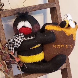 May include: A black and yellow felt bee with a red face and a black and white checkered scarf holds a brown felt honey pot with the word "Honey" embroidered on it. The bee is sitting on a wooden ladder.