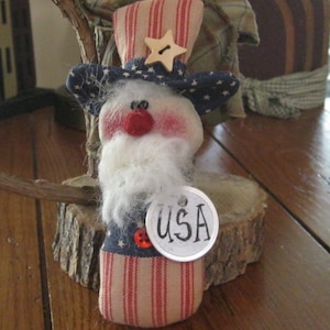May include: A handmade patriotic doll with a red, white, and blue striped hat, a white beard, and a red button nose. The doll is holding a silver tag that says "USA".