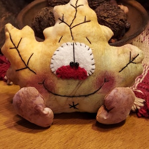 May include: A leaf-shaped plush toy with a smiling face, stitched details, and small, stuffed feet. The toy is primarily yellow with brown accents, a white felt nose, and a red mouth. The background includes a bowl of nuts.