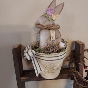 May include: A wooden Easter bunny decoration with a flower crown and a jute bow. The bunny sits in a cream-colored pot with a green and white striped ribbon. The word "HOP" is visible on a small tag. The pot is filled with faux grass and an egg.