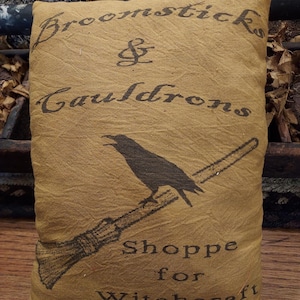 May include: A rectangular mustard yellow fabric pillow featuring black printed text that reads Broomsticks & Cauldrons Shoppe for Witchcraft 1762. The design includes a black silhouette of a crow perched on a broomstick centered on the pillow.