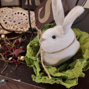 May include: A white felt bunny with black eyes and twine whiskers sits in a bed of green fabric. The bunny is surrounded by green fabric, and the twine is wrapped around the bunny's head. The bunny is a decorative Easter or spring-themed item.