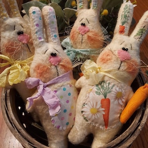 May include: Four handmade felt Easter bunny figurines in a brown wicker basket. The bunnies are white with pink noses and have different fabric accents. One bunny has a purple ribbon, another has a yellow ribbon, and another has a green and blue ribbon. The fourth bunny has a carrot.