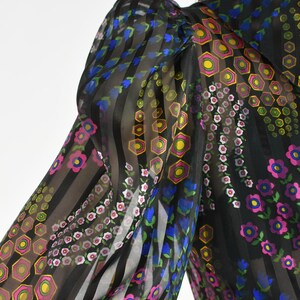 K&ouml;nnte beinhalten: Schwarze transparente Bluse mit vertikalen Streifen und floralen Mustern. Die &Auml;rmel zeigen eine Mischung aus rosa, blauen, gelben und gr&uuml;nen Blumen- und geometrischen Designs. Die Bluse hat einen hohen Kragen.