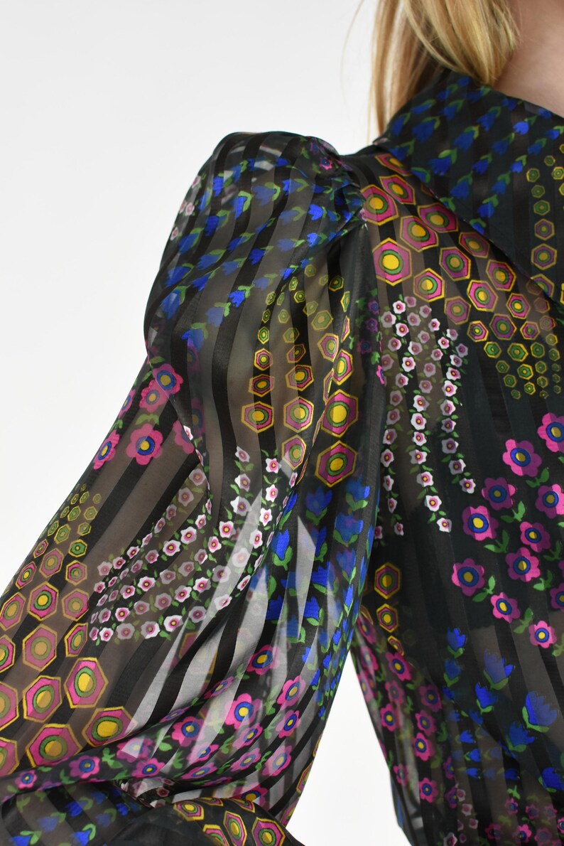 K&ouml;nnte beinhalten: Schwarze transparente Bluse mit vertikalen Streifen und floralen Mustern. Die &Auml;rmel zeigen eine Mischung aus rosa, blauen, gelben und gr&uuml;nen Blumen- und geometrischen Designs. Die Bluse hat einen hohen Kragen.