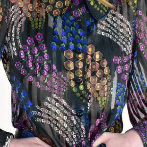 K&ouml;nnte beinhalten: Eine transparente schwarze Bluse mit einem farbenfrohen Blumenmuster. Die Bluse hat einen Kragen, lange &Auml;rmel und eine Knopfleiste. Das Blumenmuster enth&auml;lt rosa, blaue, gelbe und gr&uuml;ne Akzente. Die Bluse wird mit einer blauen Jeans und einem braunen G&uuml;rtel getragen.
