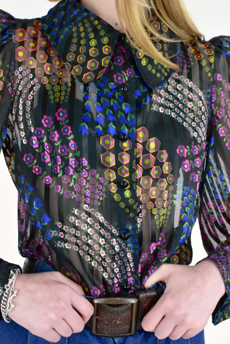 K&ouml;nnte beinhalten: Eine transparente schwarze Bluse mit einem farbenfrohen Blumenmuster. Die Bluse hat einen Kragen, lange &Auml;rmel und eine Knopfleiste. Das Blumenmuster enth&auml;lt rosa, blaue, gelbe und gr&uuml;ne Akzente. Die Bluse wird mit einer blauen Jeans und einem braunen G&uuml;rtel getragen.