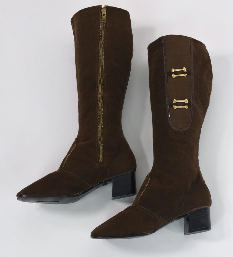 Puede incluir: Un par de botas marrones hasta la rodilla. Una bota tiene una cremallera dorada de cuerpo entero, la otra tiene dos hebillas decorativas doradas. Ambas botas tienen punta afilada y tac&oacute;n de bloque negro.