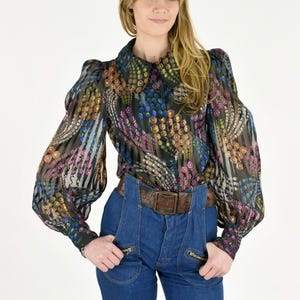 K&ouml;nnte beinhalten: Eine transparente schwarze Bluse mit farbenfrohem Blumenmuster, langen Puff&auml;rmeln und einem Kragen. Die Bluse wird mit einer hoch taillierten Jeanshose und einem braunen Lederg&uuml;rtel kombiniert.
