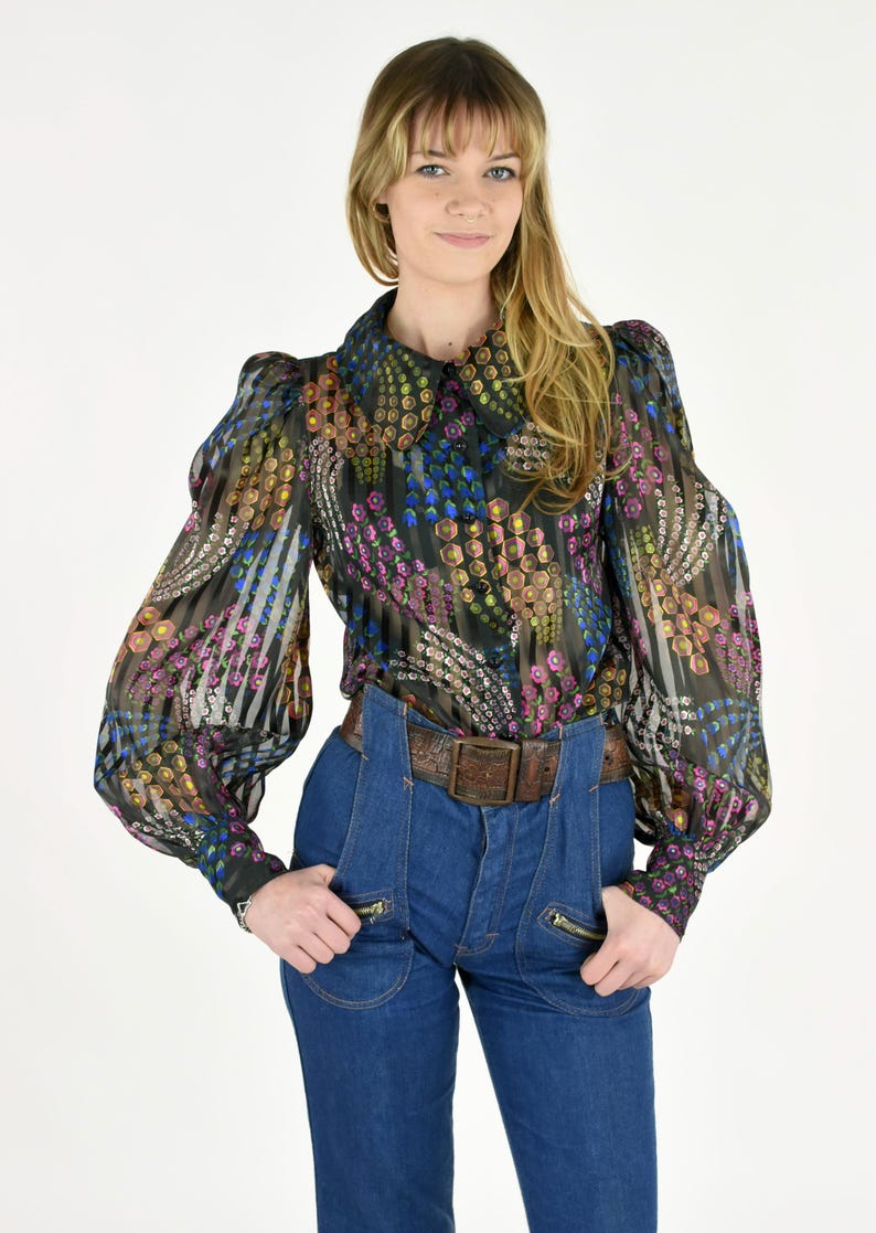 K&ouml;nnte beinhalten: Eine transparente schwarze Bluse mit farbenfrohem Blumenmuster, langen Puff&auml;rmeln und einem Kragen. Die Bluse wird mit einer hoch taillierten Jeanshose und einem braunen Lederg&uuml;rtel kombiniert.
