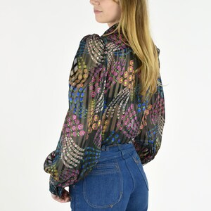 K&ouml;nnte beinhalten: Eine transparente Bluse mit langen &Auml;rmeln und einem bunten Blumenmuster in Rosa, Blau und Gelb. Dazu eine hoch taillierte, dunkelblaue Jeans. Die &Auml;rmel sind an den Handgelenken gerafft.