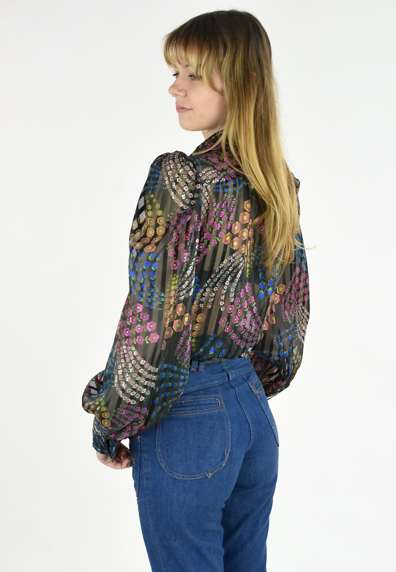 K&ouml;nnte beinhalten: Eine transparente Bluse mit langen &Auml;rmeln und einem bunten Blumenmuster in Rosa, Blau und Gelb. Dazu eine hoch taillierte, dunkelblaue Jeans. Die &Auml;rmel sind an den Handgelenken gerafft.