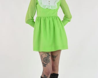 May include: A lime green mini dress with long, sheer sleeves and a white ruffled collar. The dress features a fitted bodice and a flared skirt. The model is wearing dark brown lace-up boots.