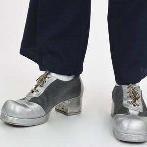 May include: A pair of lace-up shoes featuring a metallic silver toe cap and heel, contrasted with dark gray suede side panels. The shoes have a thick platform sole, a block heel, and brown and white striped laces, worn with dark navy trousers.