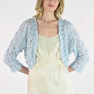 May include: A light blue, open-front bolero jacket with a floral lace design. The jacket has three-quarter sleeves and is adorned with small, white beads. The bolero is worn over a cream-colored slip dress.