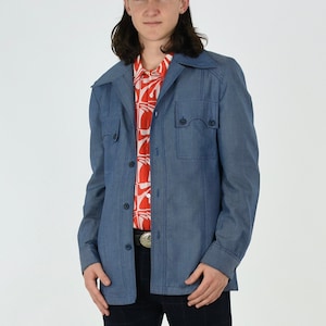 May include: A blue denim jacket with a button-up front and two flap pockets. The jacket has a collar and long sleeves. The person is wearing a red and white patterned shirt underneath.