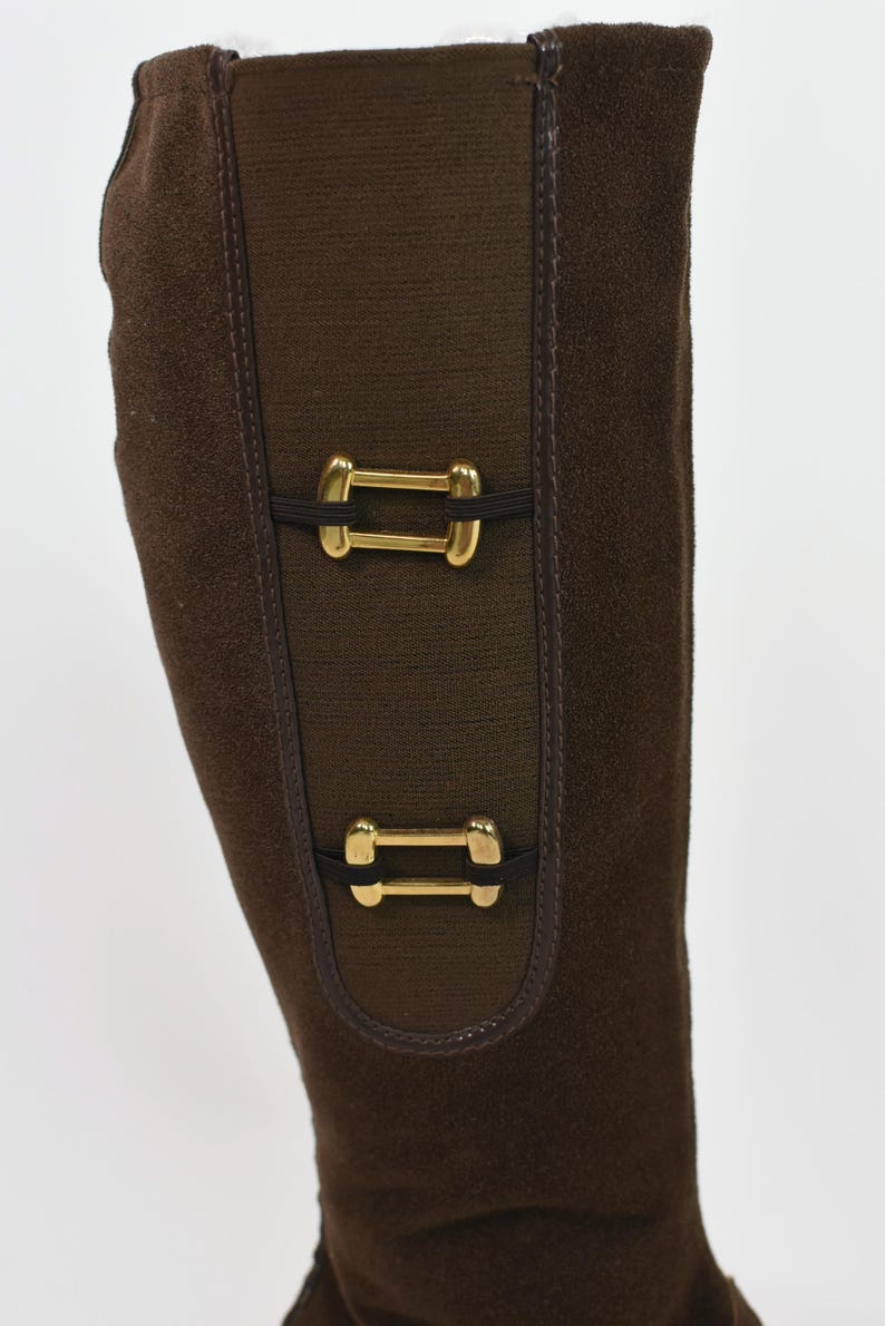 Puede incluir: Bota alta de ante marr&oacute;n con detalles dorados. La bota presenta un panel el&aacute;stico marr&oacute;n oscuro con dos hebillas rectangulares doradas. La bota est&aacute; hecha de ante y tiene un dise&ntilde;o cl&aacute;sico.