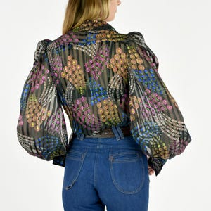 K&ouml;nnte beinhalten: Eine transparente schwarze Bluse mit einem bunten Blumenmuster und langen, puff&auml;rmeligen &Auml;rmeln. Die Bluse wird mit einer hoch taillierten, dunkelblauen Jeans kombiniert. Das Blumenmuster enth&auml;lt rosa, gelbe und blaue Akzente.