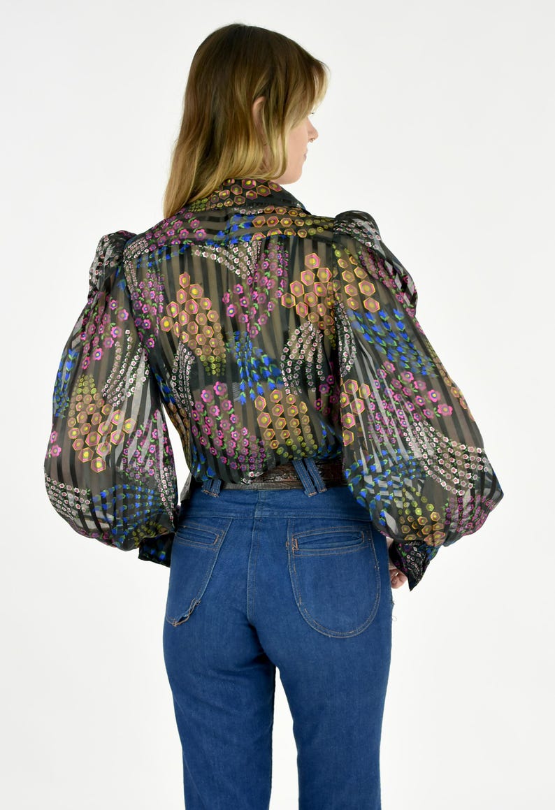 K&ouml;nnte beinhalten: Eine transparente schwarze Bluse mit einem bunten Blumenmuster und langen, puff&auml;rmeligen &Auml;rmeln. Die Bluse wird mit einer hoch taillierten, dunkelblauen Jeans kombiniert. Das Blumenmuster enth&auml;lt rosa, gelbe und blaue Akzente.