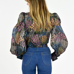K&ouml;nnte beinhalten: Eine transparente schwarze Bluse mit einem Blumenmuster in Rosa, Gelb und Blau, kombiniert mit einer hoch taillierten blauen Jeans. Die Bluse hat lange, volumin&ouml;se &Auml;rmel und eine taillierte Taille. Die Jeans hat ein klassisches F&uuml;nf-Taschen-Design.