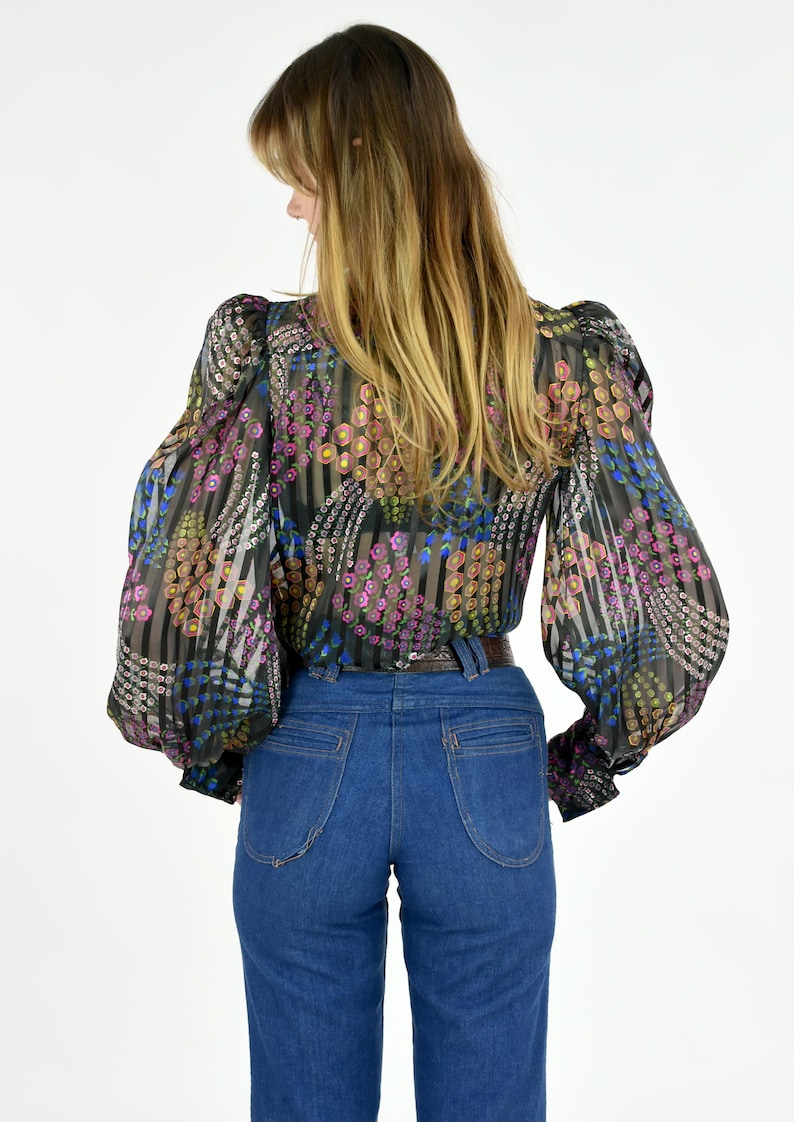 K&ouml;nnte beinhalten: Eine transparente schwarze Bluse mit einem Blumenmuster in Rosa, Gelb und Blau, kombiniert mit einer hoch taillierten blauen Jeans. Die Bluse hat lange, volumin&ouml;se &Auml;rmel und eine taillierte Taille. Die Jeans hat ein klassisches F&uuml;nf-Taschen-Design.