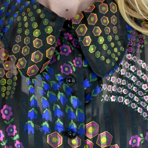 K&ouml;nnte beinhalten: Schwarze Bluse mit floralem Muster und gro&szlig;em Kragen. Der Kragen und die Bluse sind mit geometrischen und floralen Designs in Gelb, Gr&uuml;n, Pink und Blau verziert. Der Stoff ist transparent mit vertikalen Streifen.