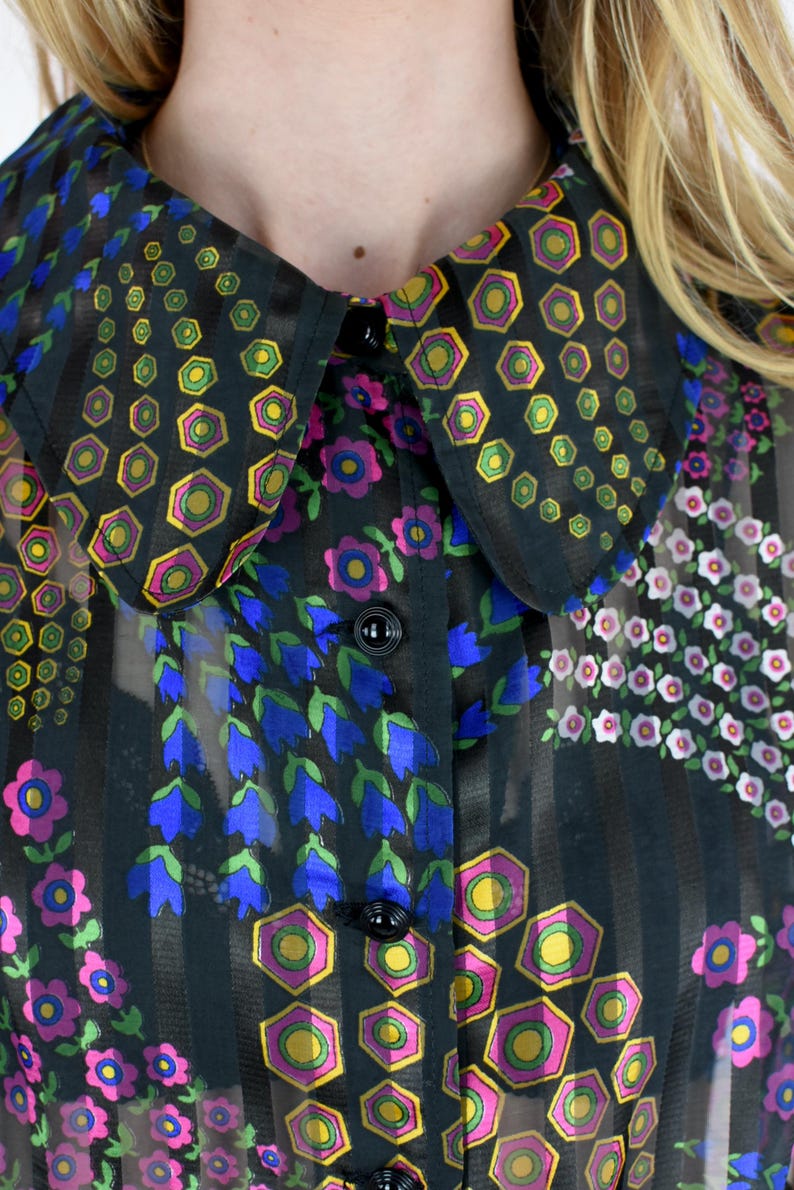 K&ouml;nnte beinhalten: Schwarze Bluse mit floralem Muster und gro&szlig;em Kragen. Der Kragen und die Bluse sind mit geometrischen und floralen Designs in Gelb, Gr&uuml;n, Pink und Blau verziert. Der Stoff ist transparent mit vertikalen Streifen.
