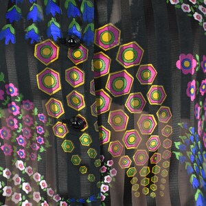 K&ouml;nnte beinhalten: Schwarze, transparente Bluse mit vertikalen Streifen und floralen Mustern. Die Bluse hat blaue glockenf&ouml;rmige Blumen, rosa und wei&szlig;e Blumen und geometrische Formen mit rosa, gelben und gr&uuml;nen Akzenten. Schwarze Kn&ouml;pfe sind sichtbar.
