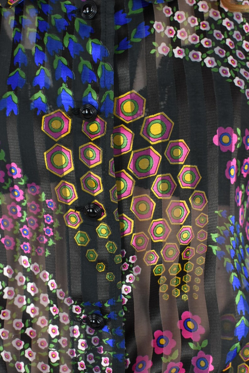 K&ouml;nnte beinhalten: Schwarze, transparente Bluse mit vertikalen Streifen und floralen Mustern. Die Bluse hat blaue glockenf&ouml;rmige Blumen, rosa und wei&szlig;e Blumen und geometrische Formen mit rosa, gelben und gr&uuml;nen Akzenten. Schwarze Kn&ouml;pfe sind sichtbar.
