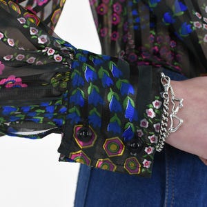 K&ouml;nnte beinhalten: Schwarze transparente Bluse mit vertikalen Streifen und Blumenmustern in Rosa, Blau und Wei&szlig;. Die Manschette zeigt blaue Glockenblumen und geometrische Formen. Ein silbernes Armband mit Sternanh&auml;nger ist sichtbar.