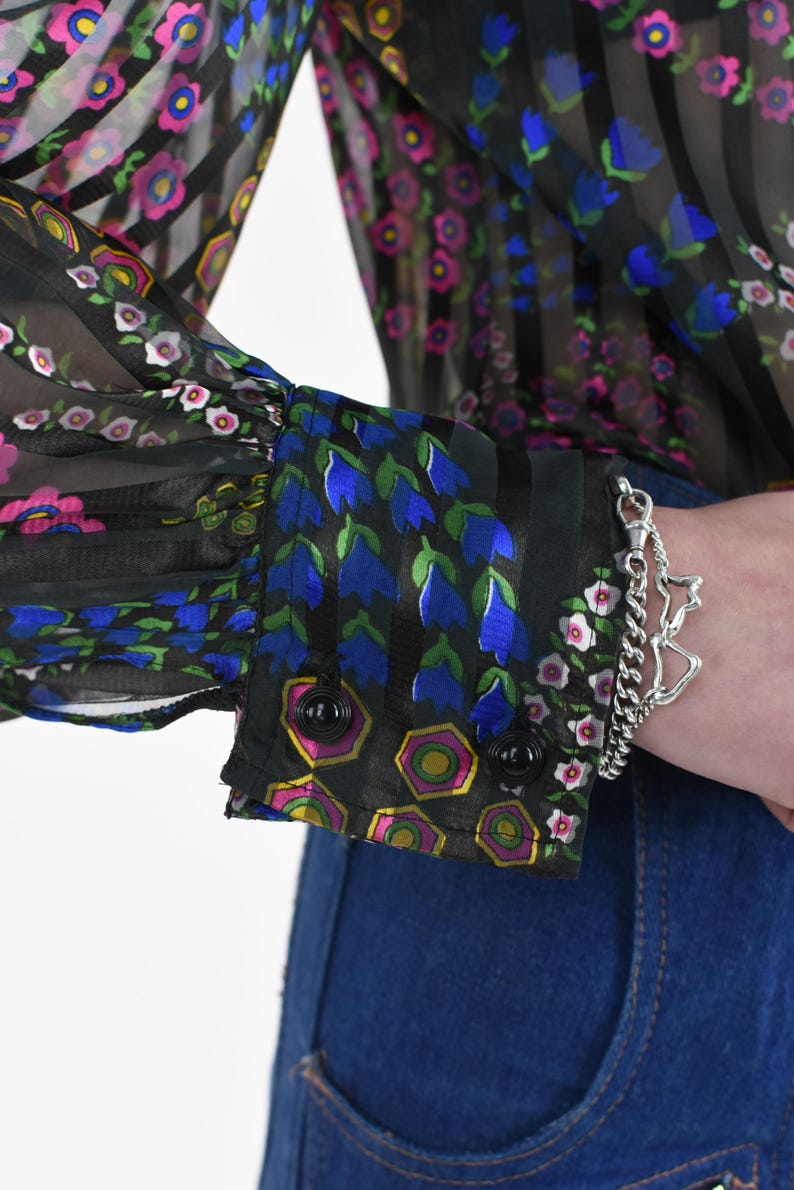 K&ouml;nnte beinhalten: Schwarze transparente Bluse mit vertikalen Streifen und Blumenmustern in Rosa, Blau und Wei&szlig;. Die Manschette zeigt blaue Glockenblumen und geometrische Formen. Ein silbernes Armband mit Sternanh&auml;nger ist sichtbar.