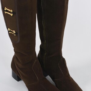 Puede incluir: Un par de botas altas de ante marr&oacute;n. Las botas tienen punta cuadrada, tac&oacute;n bajo y cremallera lateral. Adornos dorados decoran la parte superior de la ca&ntilde;a.