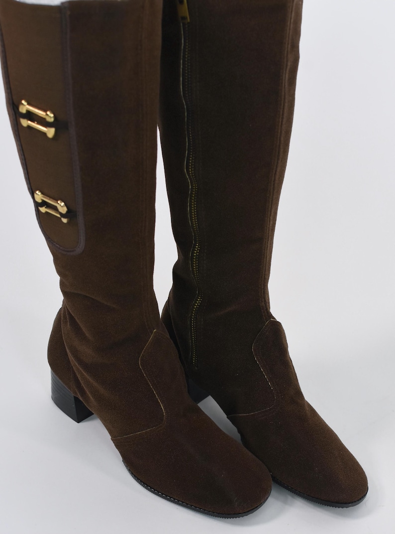 Puede incluir: Un par de botas altas de ante marr&oacute;n. Las botas tienen punta cuadrada, tac&oacute;n bajo y cremallera lateral. Adornos dorados decoran la parte superior de la ca&ntilde;a.