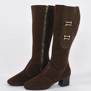 Puede incluir: Un par de botas marrones hasta la rodilla. Una bota tiene una cremallera dorada de cuerpo entero. La otra bota tiene un panel decorativo con dos hebillas doradas. Las botas tienen un tac&oacute;n peque&ntilde;o y negro y una puntera redondeada.