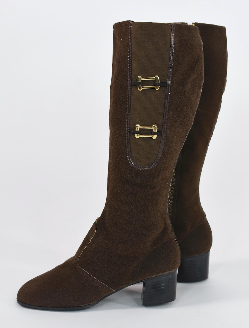 Puede incluir: Botas altas de ante marr&oacute;n con tac&oacute;n bajo. Las botas presentan un panel decorativo con dos hebillas doradas. Una cremallera recorre el lateral. Las botas est&aacute;n sobre un fondo blanco.