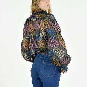 K&ouml;nnte beinhalten: Eine transparente schwarze Bluse mit farbenfrohem Blumenmuster und langen &Auml;rmeln wird mit einer hoch taillierten blauen Jeans kombiniert. Die Bluse hat ein vertikales Streifendesign mit rosa, gelben und blauen floralen Akzenten. Die Jeans ist ein klassischer Denim-Stil.