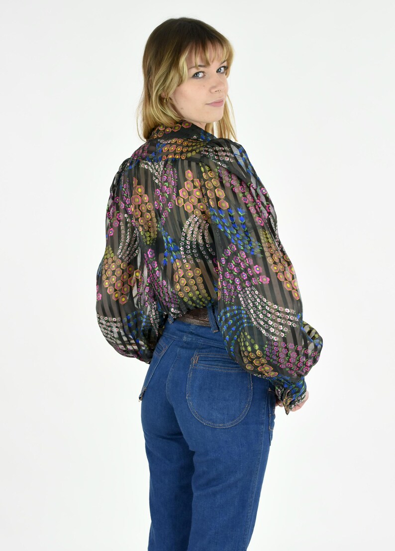 K&ouml;nnte beinhalten: Eine transparente schwarze Bluse mit farbenfrohem Blumenmuster und langen &Auml;rmeln wird mit einer hoch taillierten blauen Jeans kombiniert. Die Bluse hat ein vertikales Streifendesign mit rosa, gelben und blauen floralen Akzenten. Die Jeans ist ein klassischer Denim-Stil.