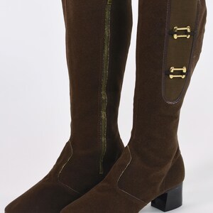 Puede incluir: Un par de botas marrones hasta la rodilla. Una bota tiene una cremallera dorada de largo completo. La otra bota tiene dos hebillas decorativas doradas. Las botas tienen un tac&oacute;n peque&ntilde;o y negro y una puntera en punta.