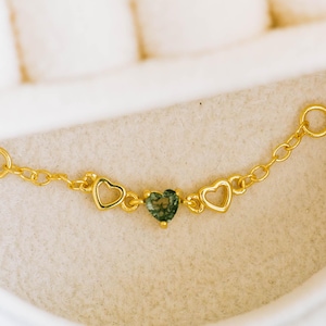 Puede incluir: Una delicada cadena dorada con tres dijes en forma de corazón y una piedra preciosa verde central. La cadena tiene pequeños bucles en cada extremo, y los dijes de corazón están espaciados uniformemente.