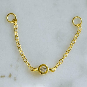 May include: A delicate gold-colored chain with a small, clear gemstone in the center. The chain has a small loop at each end. The jewelry is displayed on a white and gray marble surface.