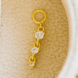 May include: A gold-colored charm with three clear, round gemstones. The charm is suspended from a small ring, and the gemstones are connected by delicate gold links. The charm is set against a soft, off-white background.