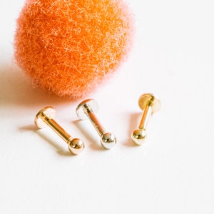 May include: Three gold and silver body jewelry pieces, each with a small ball at the end. The jewelry is arranged in a row on a white surface.