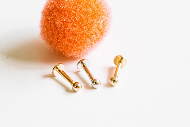 May include: Three gold and silver body jewelry pieces, each with a small ball at the end. The jewelry is arranged in a row on a white surface.
