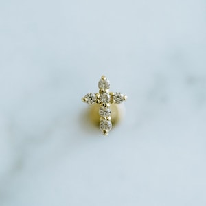May include: A small, gold-colored cross pendant with multiple clear, sparkling gemstones. The cross is set against a soft, neutral background, highlighting the jewelry's delicate design. The gemstones are arranged to form the cross shape.