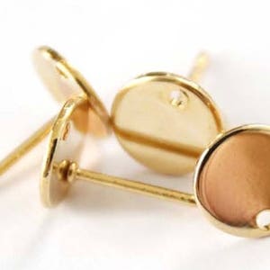 May include: Three gold-toned earring settings with a round, flat base and a post for attaching earrings. The settings have a small hole in the center for inserting a stone or other embellishment.